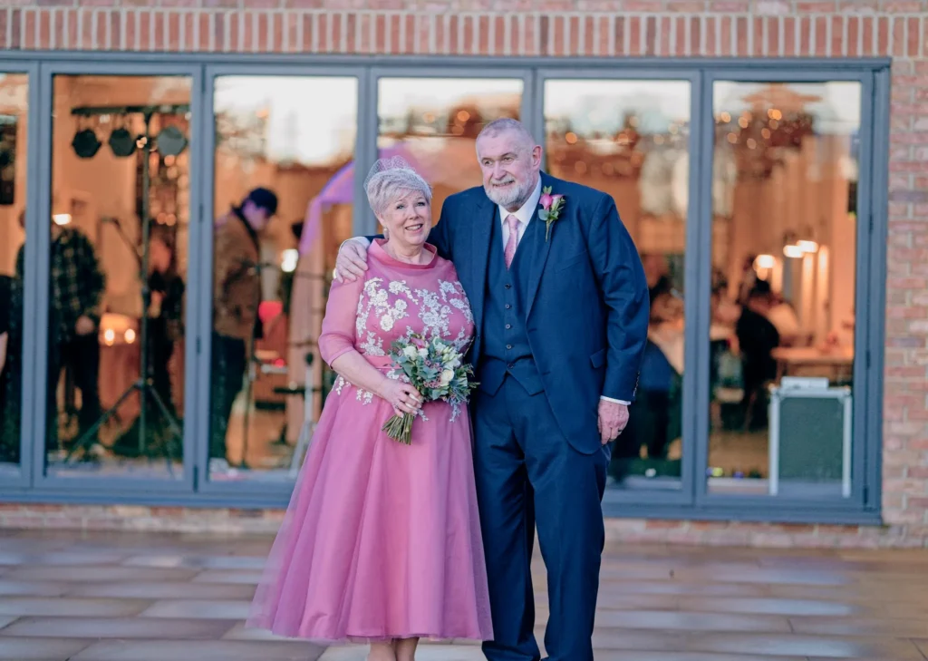 The bride in a pink dress with white lace flowers and the groom in a navy suit, his arm around her shoulders, outside a brick venue