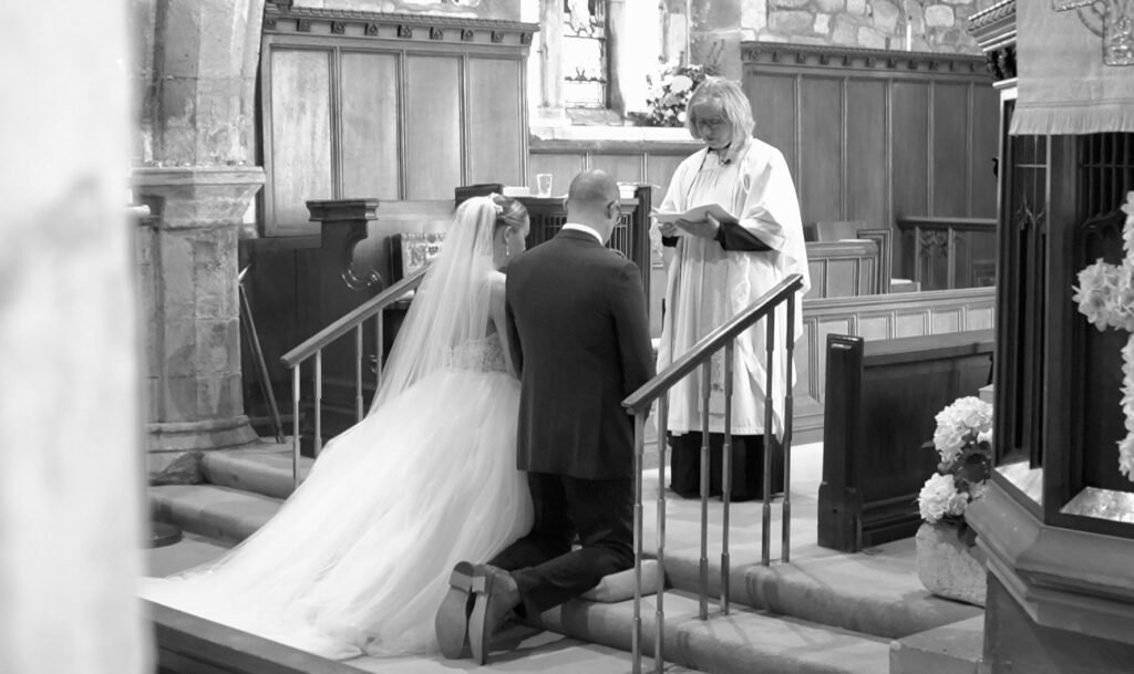 Bride and groom kneel at the altar for a blessing, in black and white