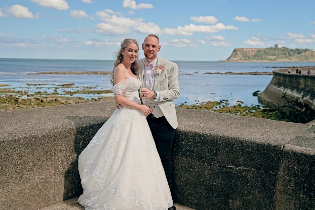 Bride and groom on a sea wall at low tide, a castle headland across the bay