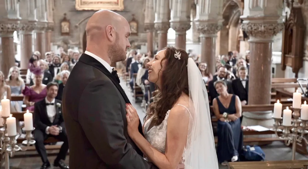 Bride and groom smile at each other in a church, candles and guests behind them