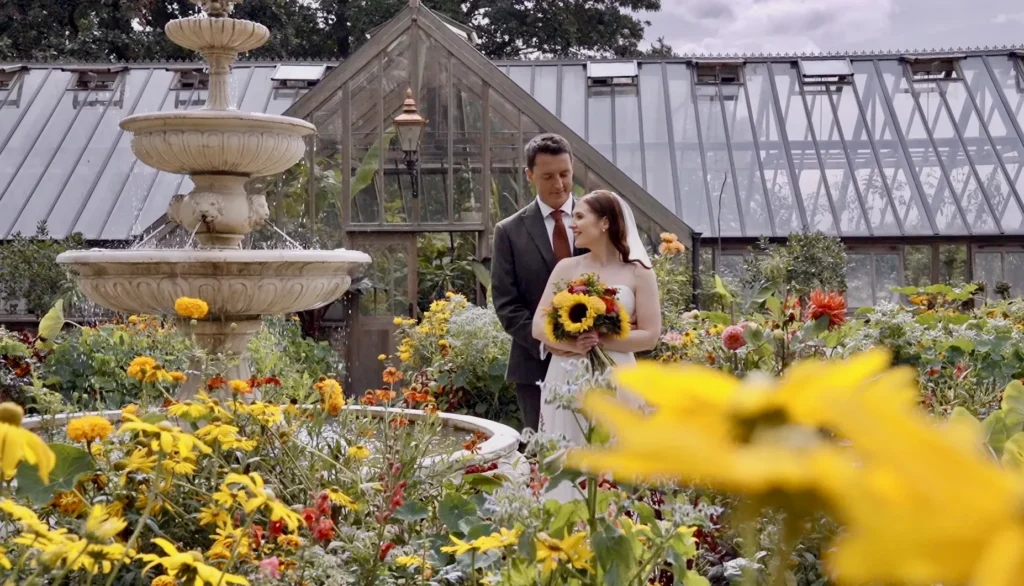 Bride and groom with a sunflower bouquet among yellow flowers, a stone fountain and a glasshouse behind