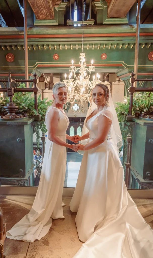 Two brides smile beneath a crystal chandelier among green Victorian ironwork
