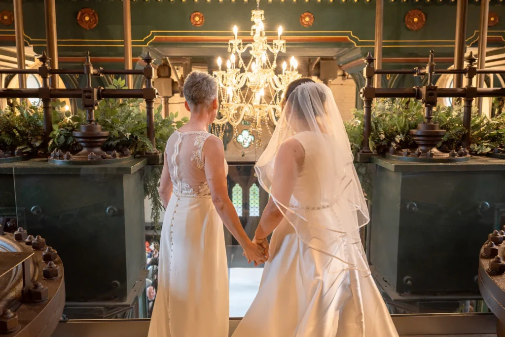 Two brides hold hands on a balcony beneath a crystal chandelier, seen from behind