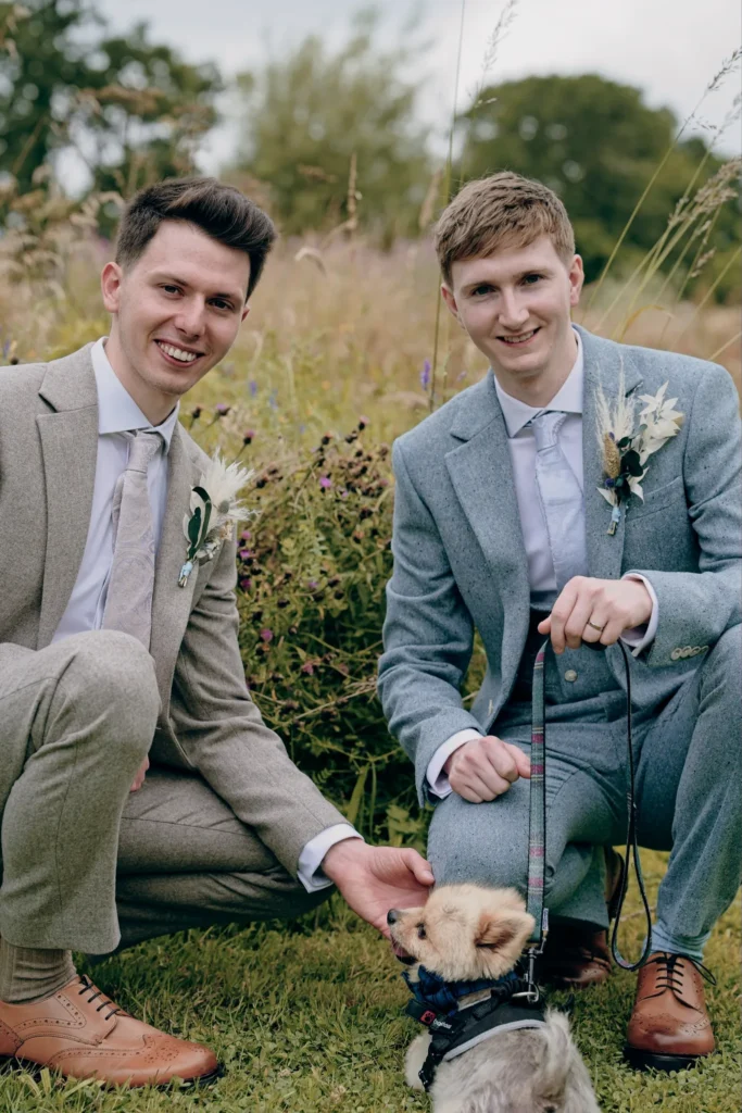 Two grooms kneel in a wild-flower meadow with their small dog