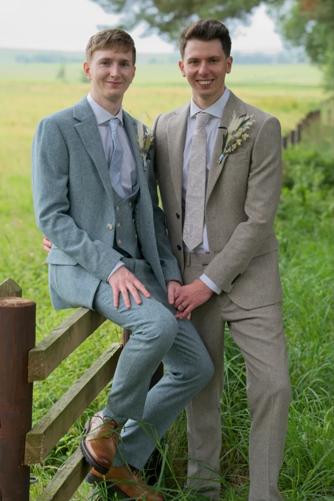 Two grooms by a wooden field fence, open fields behind
