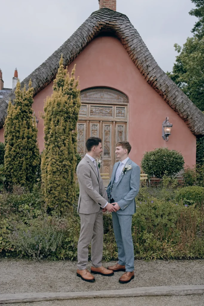 Two grooms hold hands in front of a pink cottage with a carved wooden door