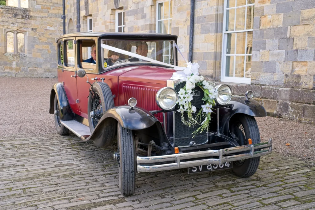 A vintage burgundy wedding car with white ribbons and lilies on a cobbled courtyard