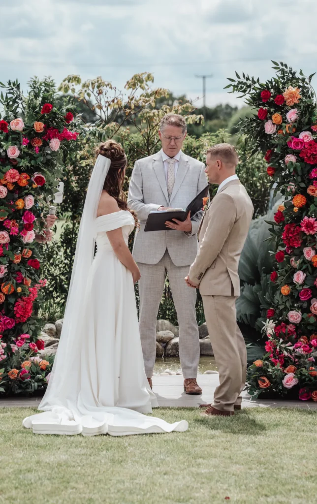 Bride and groom face the celebrant between two pillars of pink, red and orange flowers