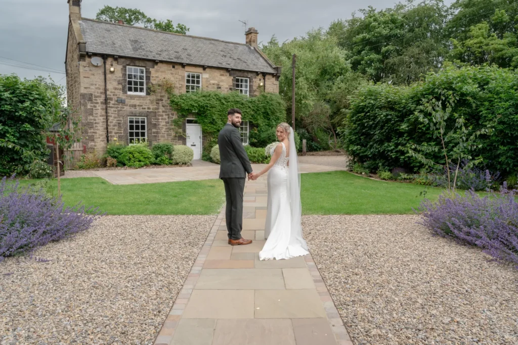 Bride and groom look back as they walk hand in hand towards a stone farmhouse