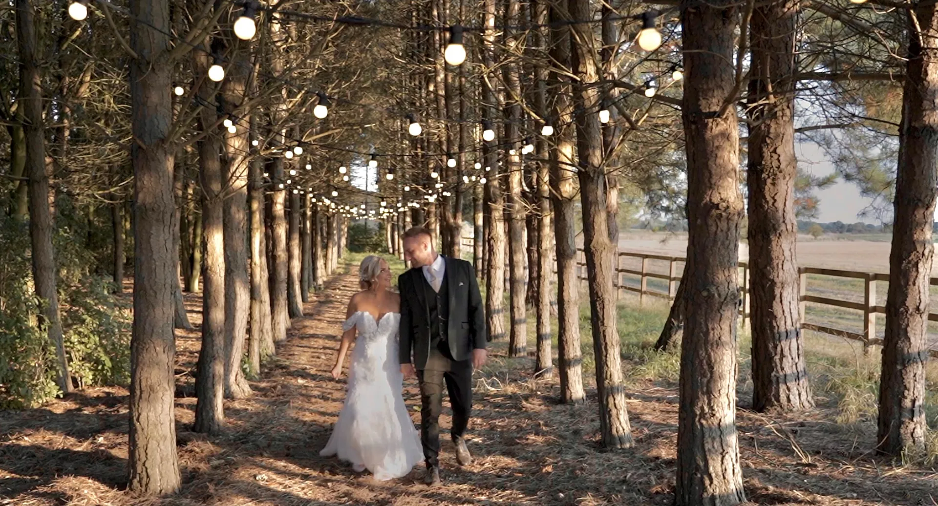 Bride and groom walk under festoon lights in an avenue of pines
